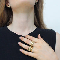 Woman wearing gold jewelry including earrings, rings, and a necklace on a plain background