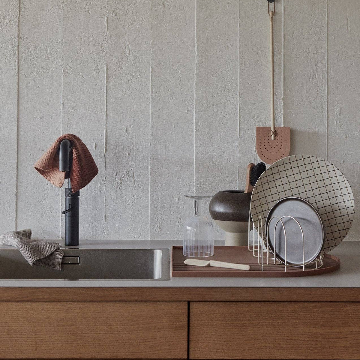 A styled kitchen countertop scene with a sink, black faucet, hanging towel, dish rack with plates, and a textured off-white paneled wall background.