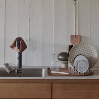 A styled kitchen countertop scene with a sink, black faucet, hanging towel, dish rack with plates, and a textured off-white paneled wall background.