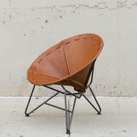 A mid-century modern leather chair with a warm tan leather seat and a black metal tripod base, set against a textured concrete wall.