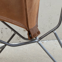 Close-up detail of a brown leather chair sling attached to a dark metal frame, showing the stitching and attachment point against a concrete wall.