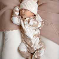 Newborn baby in a camouflage outfit and white knit hat with pom-pom on a soft pink blanket.