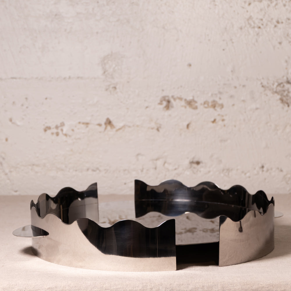A polished silver metal ring with a wavy scalloped edge, displayed on a textured off-white surface against a rustic, distressed light gray wall.