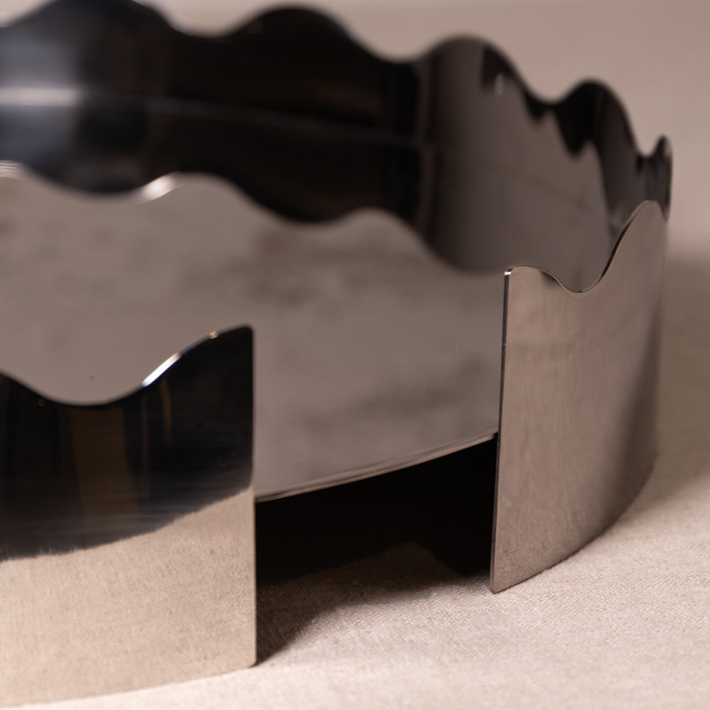 A close-up of a metallic silver wavy-edged container or tray, showing its reflective polished surface and geometric design on a neutral beige background.