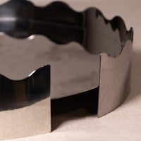 A close-up of a metallic silver wavy-edged container or tray, showing its reflective polished surface and geometric design on a neutral beige background.