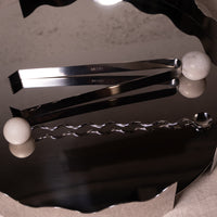 A set of polished stainless steel bar tools, including a pair of tongs and a wavy spoon, displayed on a dark, reflective surface with white marble spheres as props.