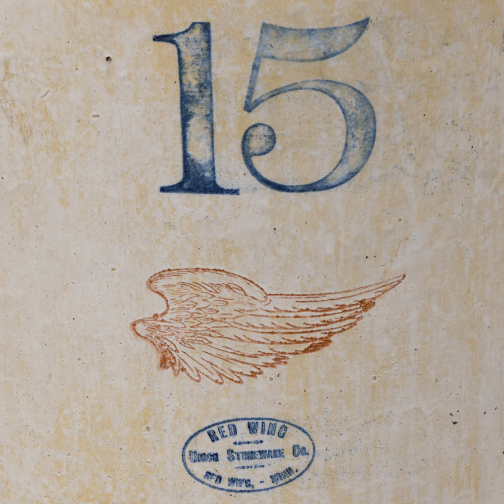 Vintage-style printed artwork or label showing the number 15 in blue at the top, a brown wing illustration in the middle, and a blue oval stamp reading Red Wing Cross Stencil Co. at the bottom on a textured beige background.