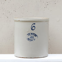 A vintage white stoneware crock pot with blue printed markings, including a blue number 6 and a circular logo for 'The Western Denver Pottery', set against a textured off-white background.