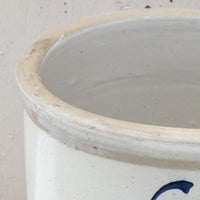 Close-up of a ceramic stoneware container, showing the interior rim with a rustic, slightly stained beige edge and a speckled off-white glaze. A partial dark blue painted letter is visible on the lower right side.