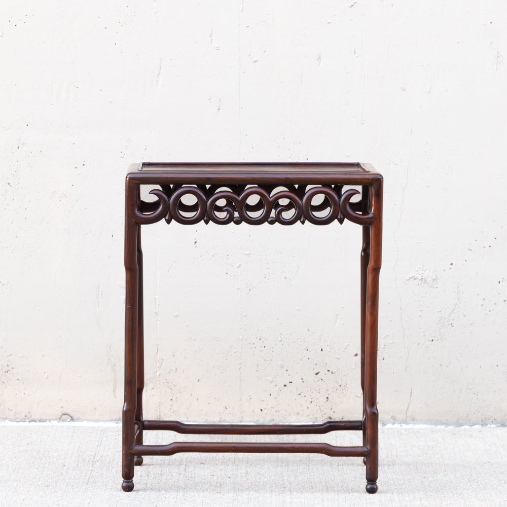 A dark brown wooden side table with a carved circular pattern along the apron, photographed against a textured off-white wall.