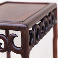 Close-up of a dark brown wooden side table with carved scrollwork legs and a rectangular top, photographed against a plain light background.
