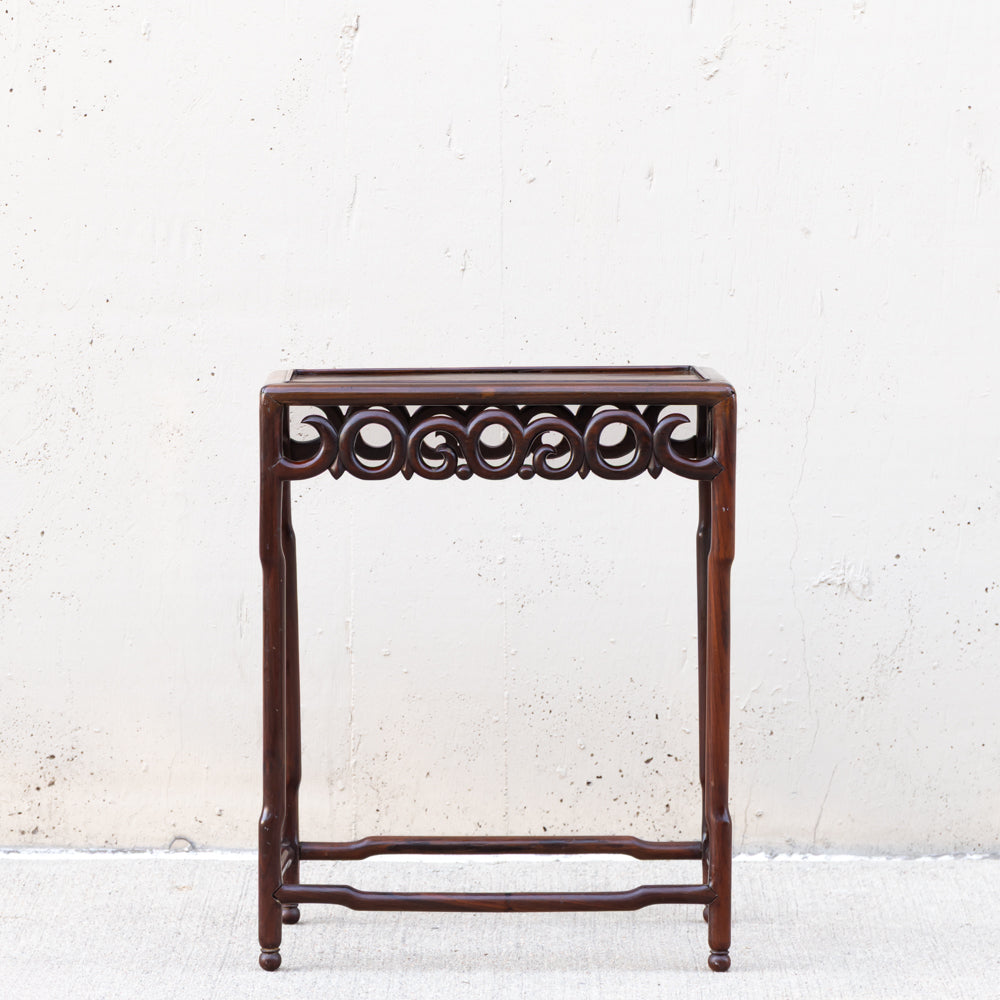A dark brown wooden side table or stand with an ornate carved scrollwork apron and open lower support, photographed against a light textured wall.