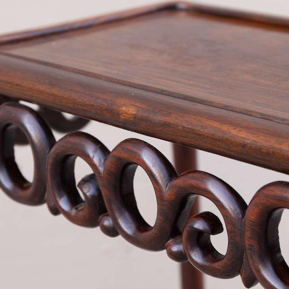 Close-up of a dark brown wooden table or stand with a smooth rectangular top and decorative carved circular scroll supports underneath, shown against a plain light background.