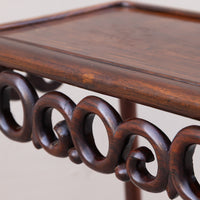 Close-up of a dark brown wooden table or stand with a smooth rectangular top and decorative carved circular scroll supports underneath, shown against a plain light background.