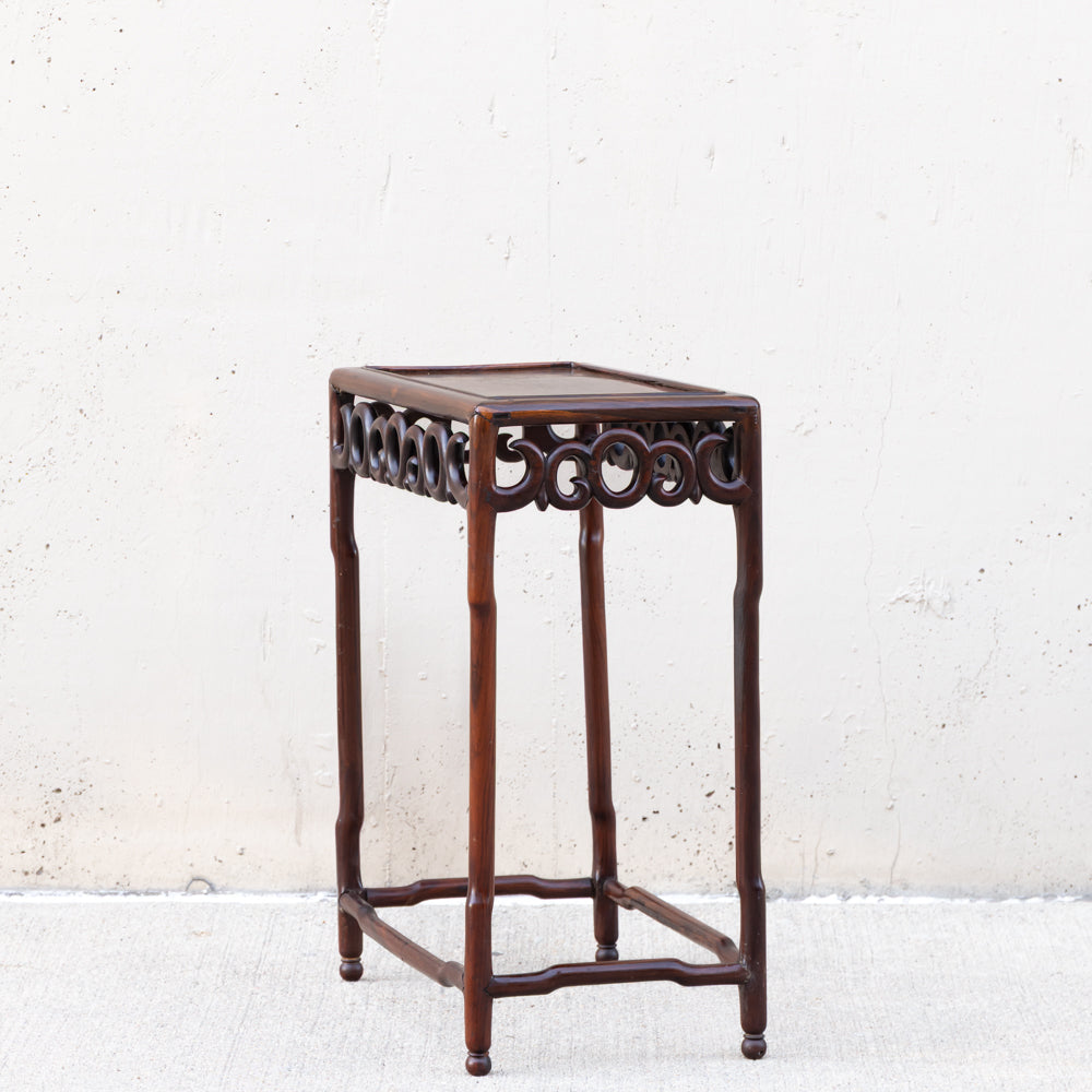 A dark brown wooden side table or stool with a rectangular top, decorative carved apron, and four slender legs, photographed against a light textured wall.