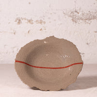 A handmade ceramic bowl with a rough, organic scalloped rim in a speckled beige-gray glaze, featuring a single thin red stripe running horizontally across the center, set against a textured off-white wall.
