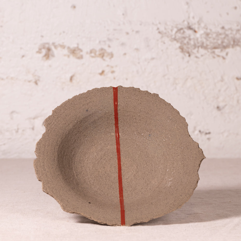 A handmade, rustic ceramic bowl with a textured, earthy brown surface and a distinctive wavy rim, featuring a thin red vertical line down the center. The bowl is photographed against a textured, off-white plaster wall.