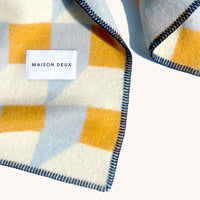 A close-up of a Maison Deux plaid blanket or scarf in cream, mustard yellow, and light blue, featuring a white brand label and dark blue stitched edges.