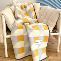 A geometric patterned throw blanket draped over a cream-colored armchair in a modern room setting with a light wood floor and a blue accent wall.