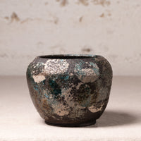 A small handmade ceramic pot with a distressed, textured finish featuring a mottled pattern of dark brown, teal, and beige glaze, set against a neutral, light textured background.