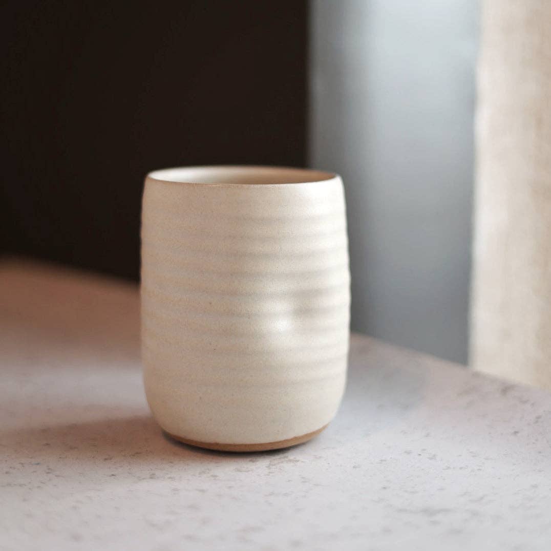 A small cylindrical ceramic cup with a textured, ribbed surface and a natural unglazed base, sitting on a light stone surface in a softly lit interior setting.