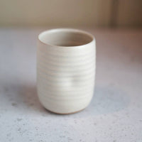 A small cylindrical ceramic cup with a textured, ribbed surface in a warm off-white or cream glaze, set on a light gray speckled countertop.