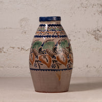 A handmade ceramic vase with a rustic, hand-painted floral pattern in blue, green, and ochre brown on a beige background, set against a textured off-white wall.