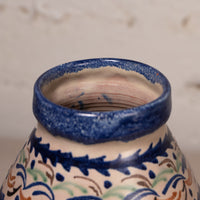 Close-up of a ceramic vase or pot showing the glazed rim and upper body, decorated with a cream base and blue, green, and brown floral scroll patterns.