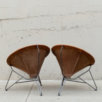Two vintage-style leather armchairs with curved brown leather seats and thin metal legs, photographed side by side against a light concrete wall.