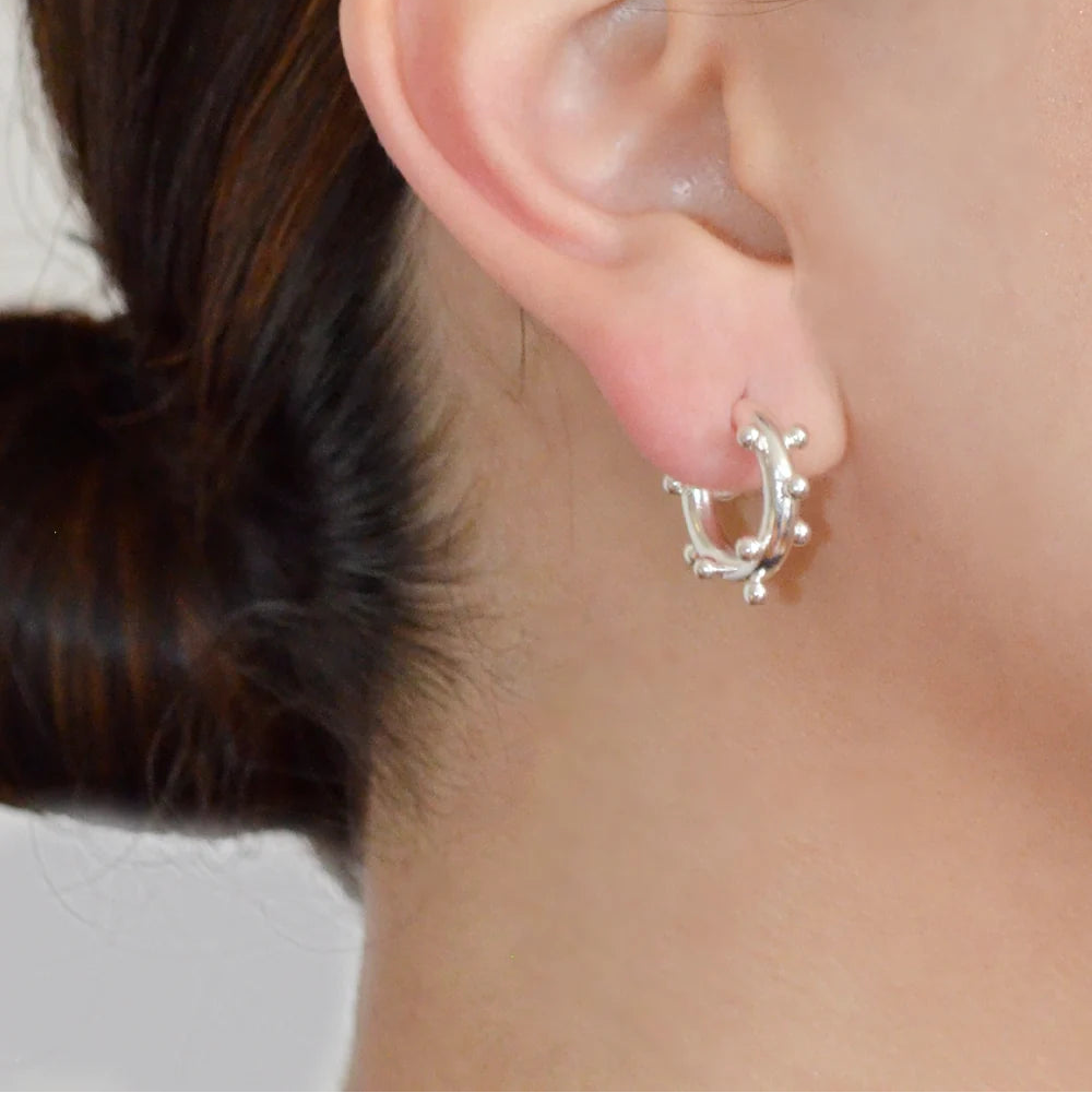 A close-up of a person's ear wearing a small silver huggie hoop earring with delicate dangling leaf-like details.