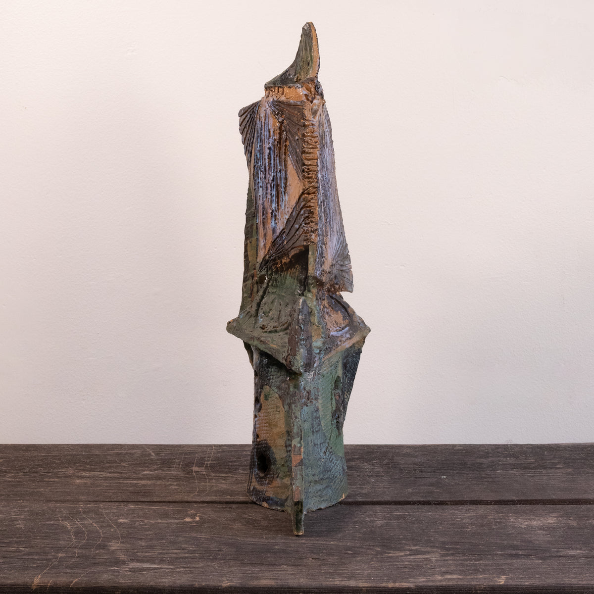 A rustic, abstract hand-carved wooden sculpture with a weathered, distressed finish in shades of brown, green, and blue, standing on a dark wooden surface against a plain off-white wall.