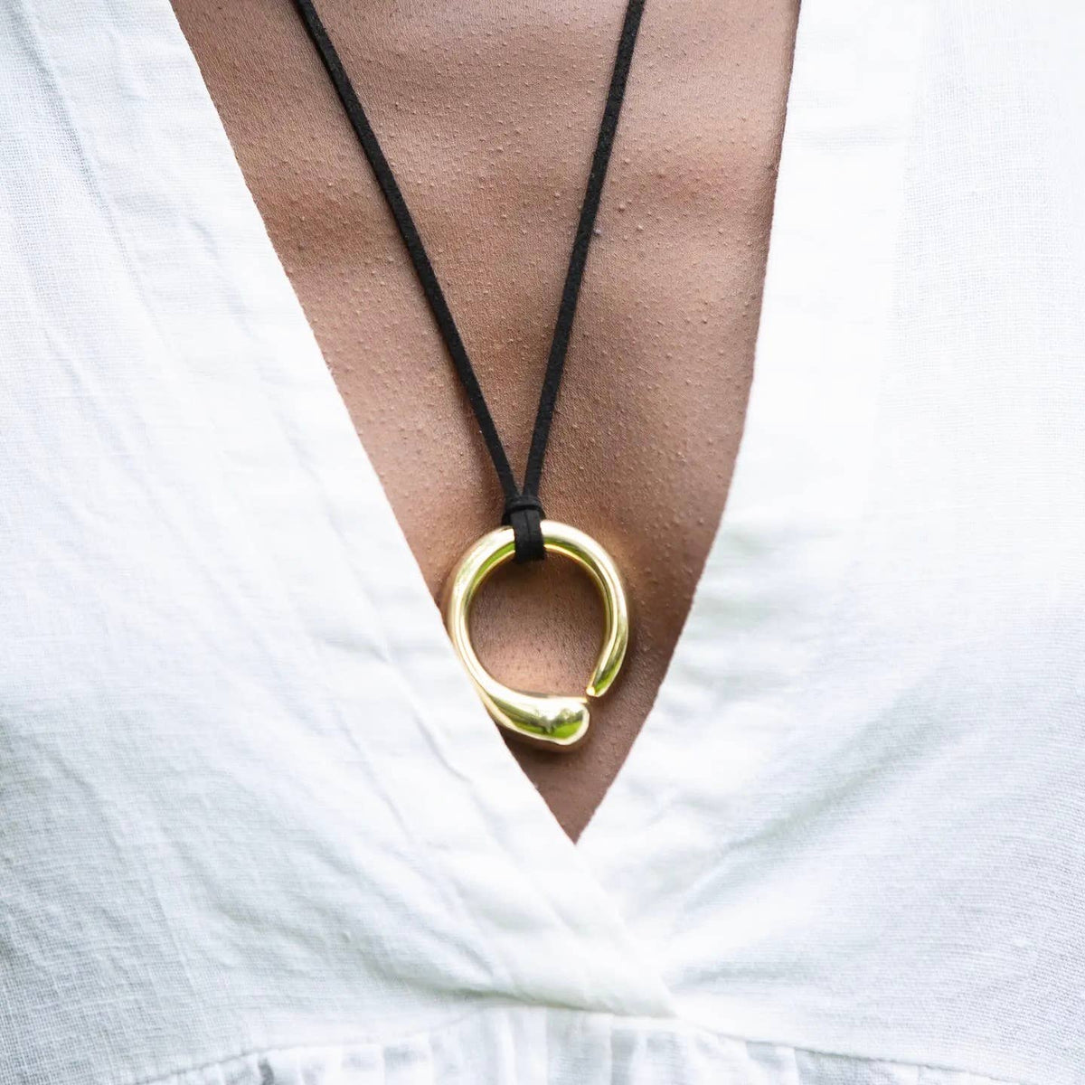 Gold necklace with a large ring pendant on a black cord, worn by a person against a white background.