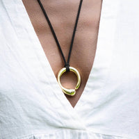 Gold necklace with a large ring pendant on a black cord, worn by a person against a white background.