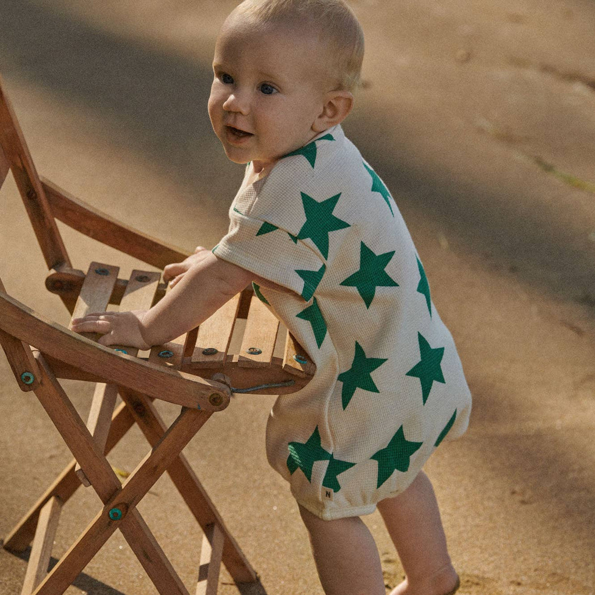 Baby wearing a white outfit with green stars, standing next to a wooden chair on a sandy surface.