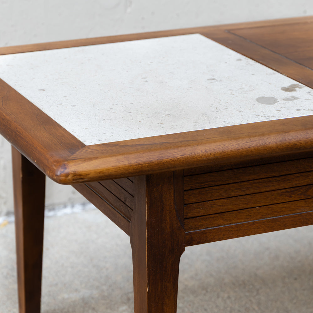 Close-up of a wooden table corner with a light beige stone tile inset on the tabletop and medium brown wood frame, shown against a neutral concrete background.