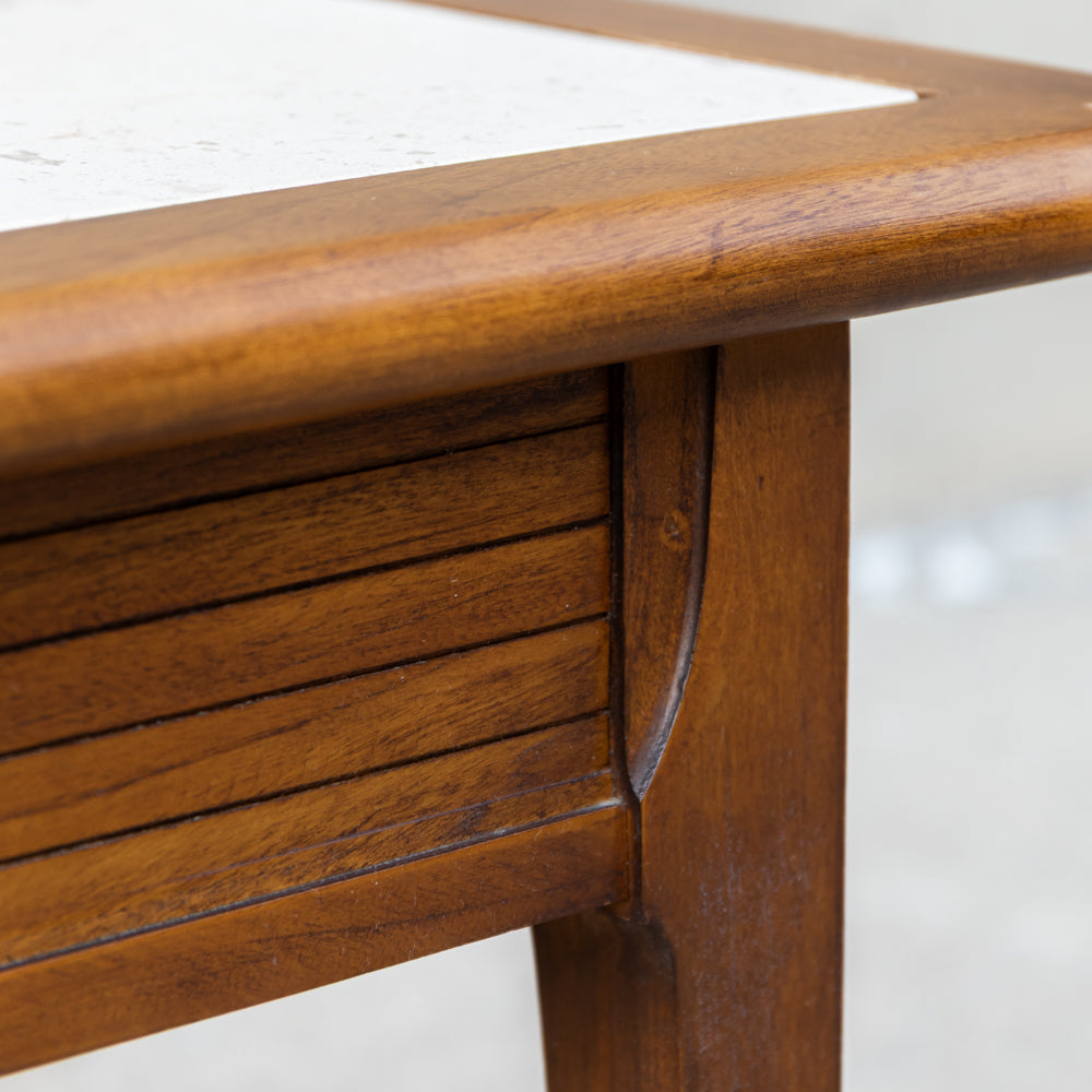 Close-up of a wooden table corner showing a warm medium-brown wood frame, a slatted drawer front, and part of a light stone or white top surface.
