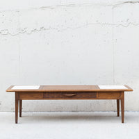 A mid-century modern wooden coffee table with a warm medium-brown wood finish, featuring a central drawer with a curved handle and two white inset panels on the top surface, set against a textured concrete wall.