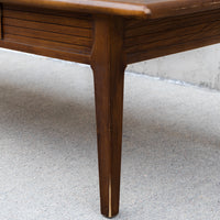 Close-up of a mid-century modern wooden table leg with a tapered design and a slatted drawer front, set on a gray concrete floor.
