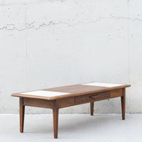A mid-century modern wooden coffee table with a medium brown wood finish, featuring a single drawer with a geometric patterned front and two light beige rectangular inlays on the top surface, set against a textured concrete wall.