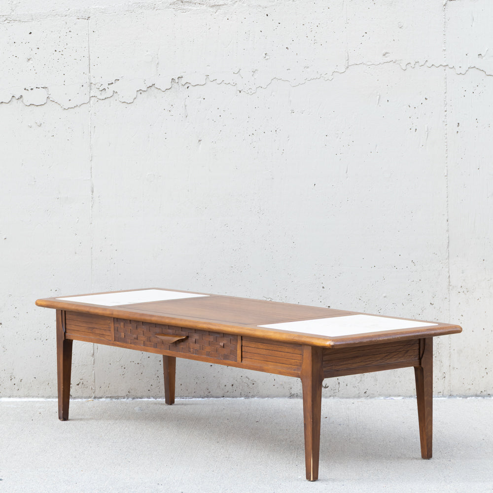 A mid-century modern wooden coffee table with a warm brown finish, featuring a single drawer with round knobs and two white rectangular inlays on the top surface, set against a textured concrete wall.