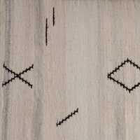 A close-up of a woven textile or rug in a warm off-white/cream tone with irregular black stitched geometric motifs, including dashes, an X, and a diamond.