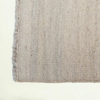 Close-up of a woven rug or textile corner showing a textured beige and light gray pattern with fringed edges on a plain white background.