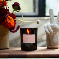 Candle with a pink label on a wooden surface next to flowers and a ceramic rabbit