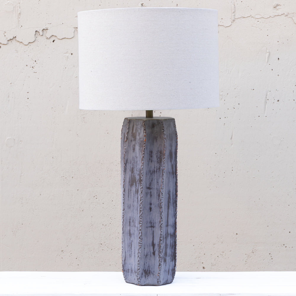 A table lamp with a cylindrical white fabric drum shade and a textured, weathered gray-blue wooden base, set against a light concrete wall.