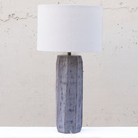 A table lamp with a textured, distressed gray-blue ceramic base and a white drum shade, set against a light beige concrete wall.