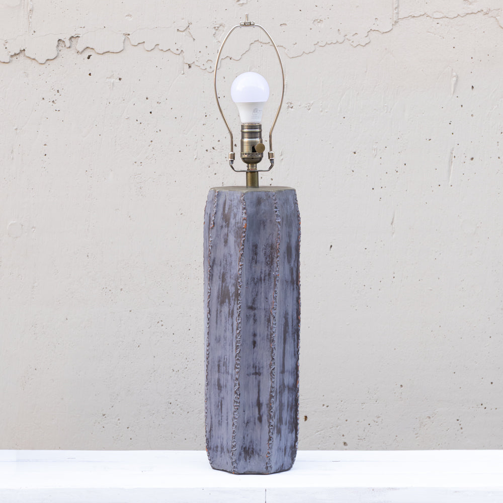 A rustic table lamp with a distressed gray-blue wooden base and a brass-toned metal socket holding a white bulb, set against a textured off-white wall.