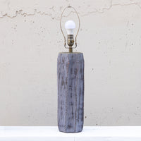 A rustic table lamp with a distressed gray-blue wooden base and a brass-toned metal socket holding a white bulb, set against a textured off-white wall.