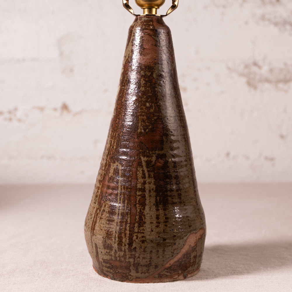 A rustic ceramic table lamp featuring a tapered, conical base with a glossy, textured brown glaze and a brass finial at the top, set against a light beige brick wall background.