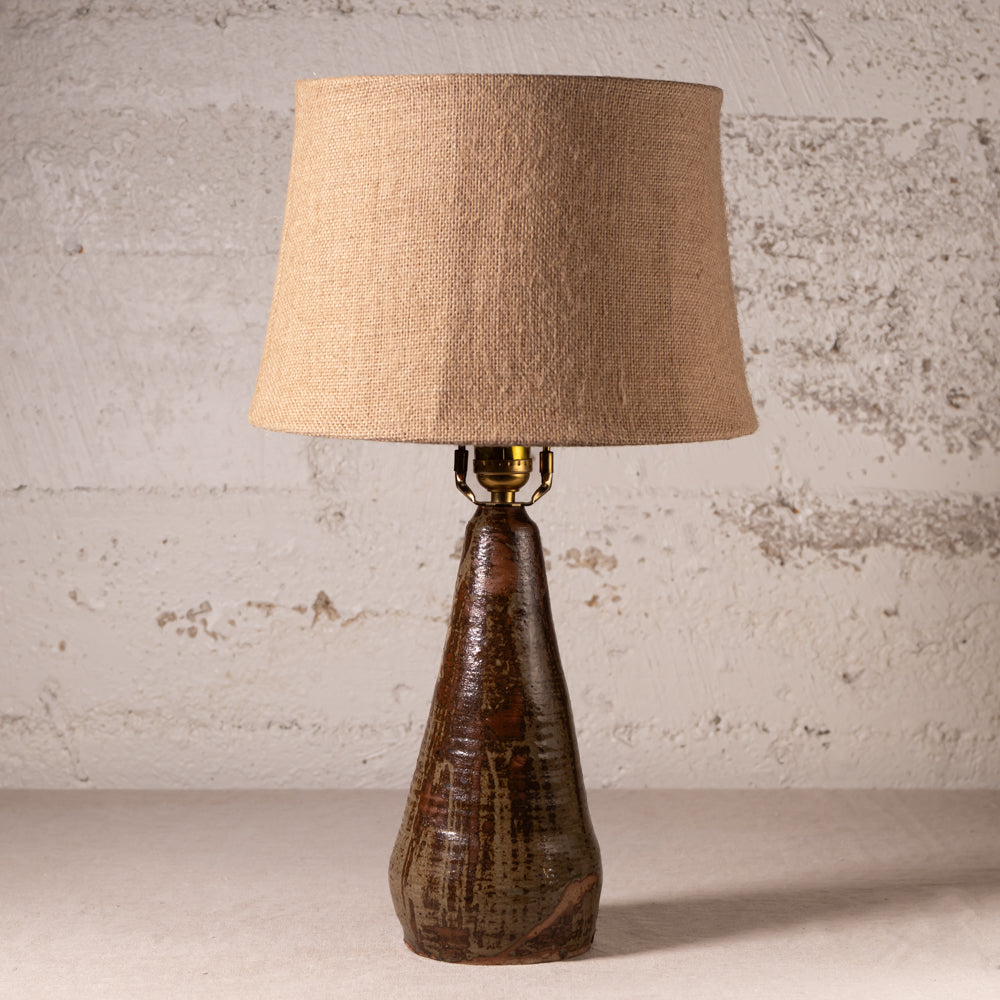 A rustic table lamp with a textured beige burlap drum shade and a dark brown glazed ceramic base, set against a distressed white brick wall.