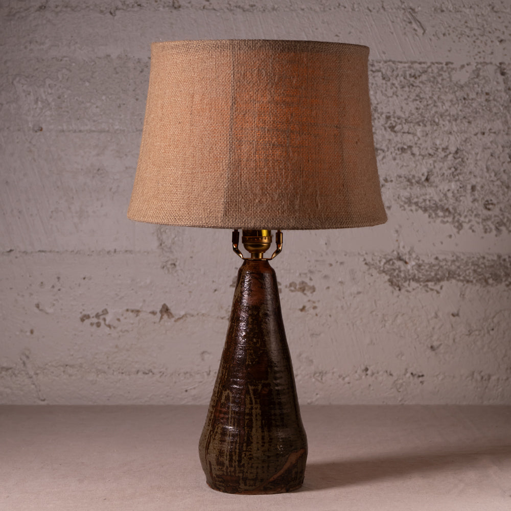 A rustic table lamp with a textured brown ceramic base and a natural beige linen drum shade, set against a distressed concrete wall.